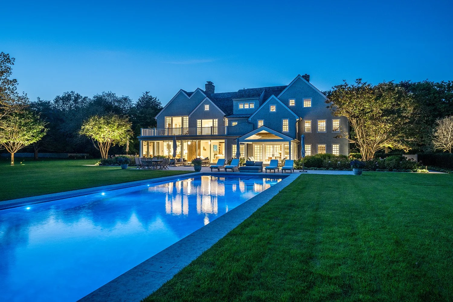 A large, well-lit house at night with a pool in the backyard. The house has multiple stories, large windows, and outdoor seating with lounge chairs and umbrellas. The yard is landscaped with trees and bushes, and the pool has blue lighting.