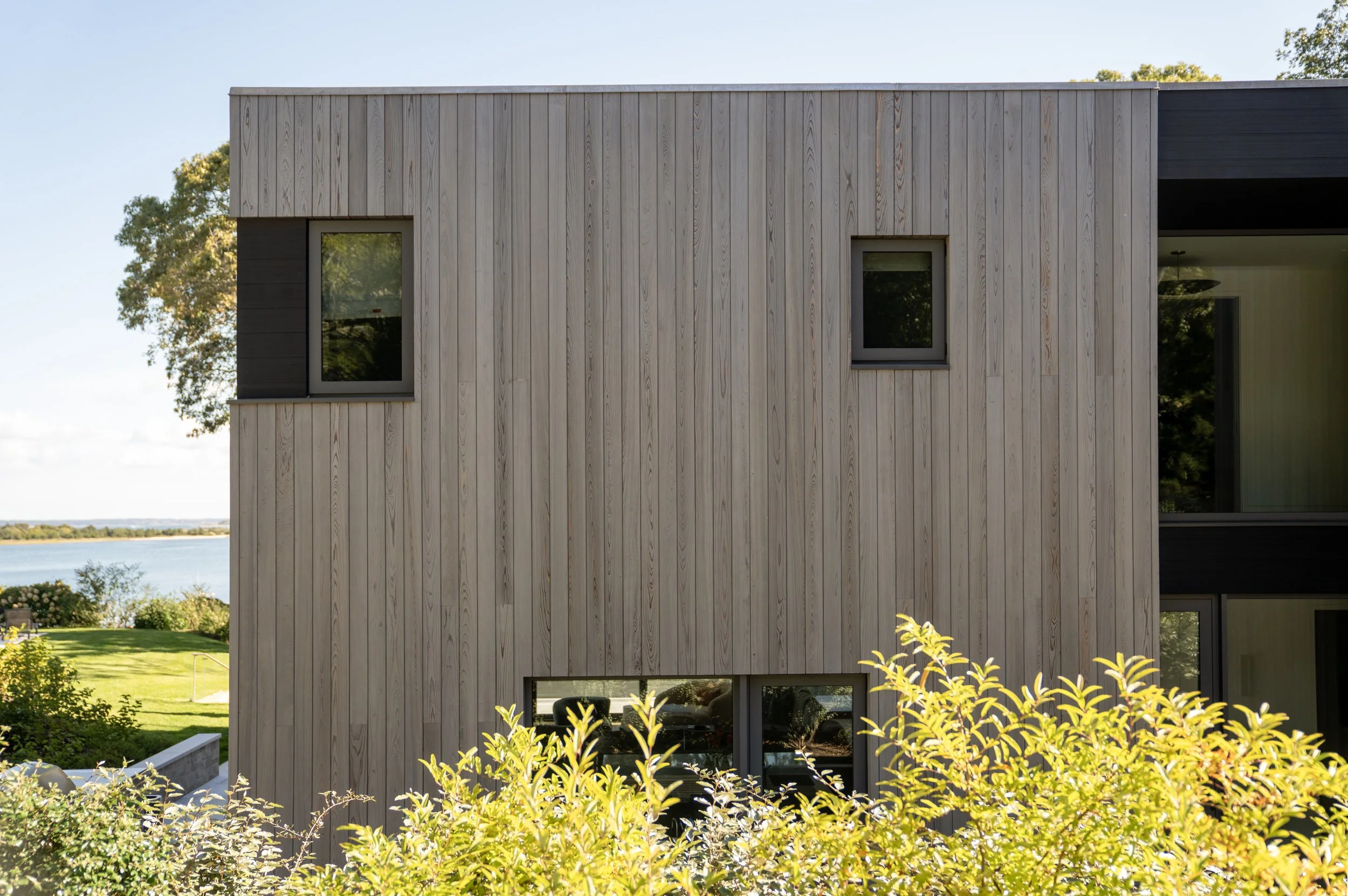 Modern building with wooden exterior, black window frames, and lush greenery in the foreground, overlooking a body of water and trees in the background.