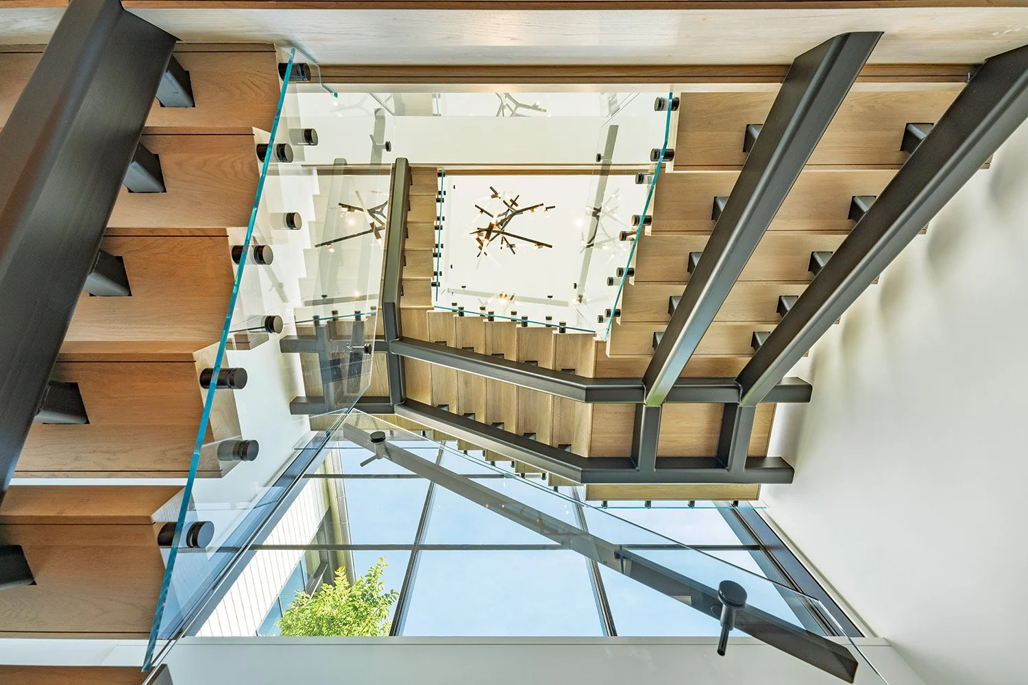 Improperly oriented photo of a modern staircase with glass railings and wooden steps, viewed from above, with large windows and a wall clock at the top.