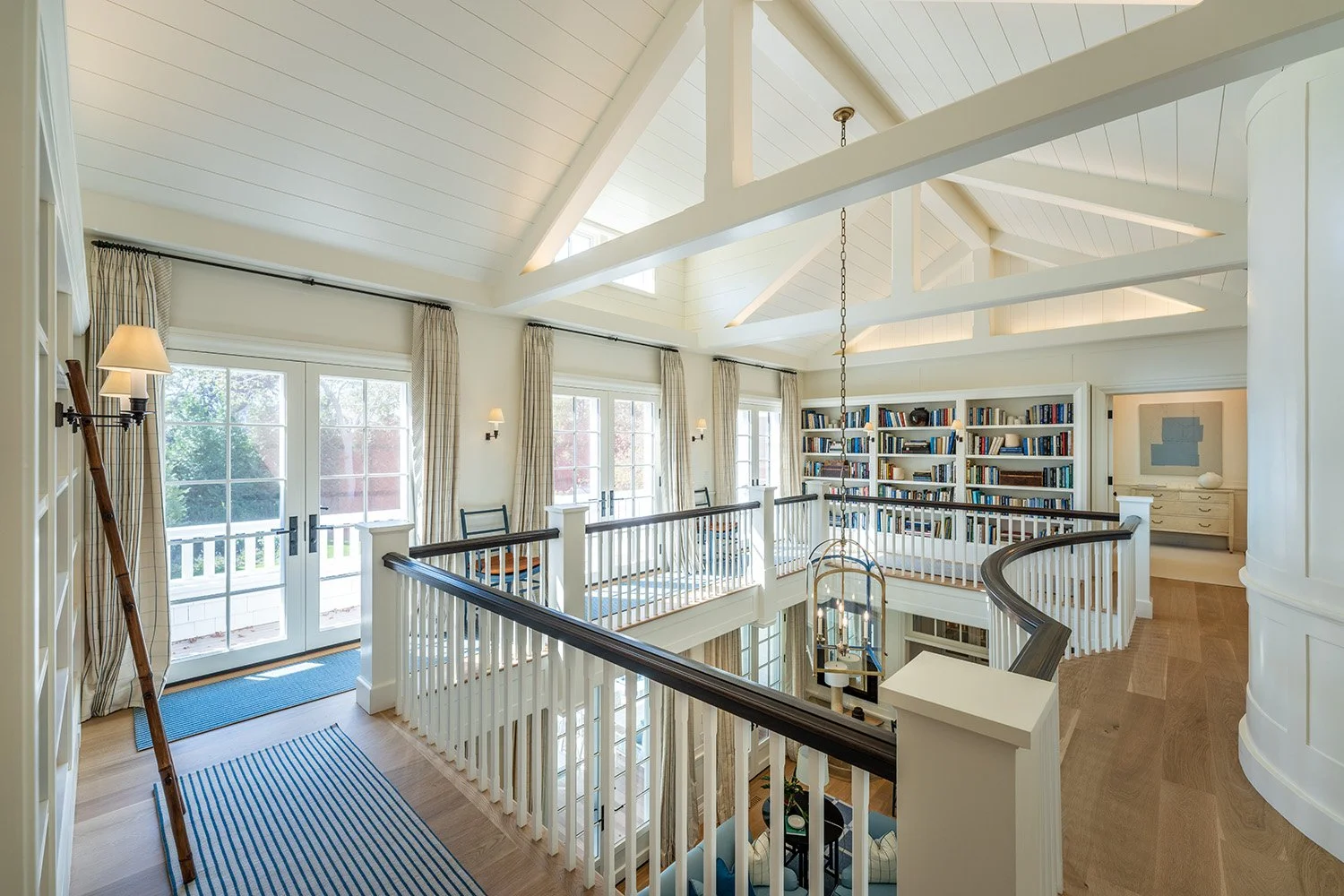 Interior view of a bright, upper-story room with large windows, white walls, and wooden flooring. The space includes a staircase with a curved handrail, a bookshelf filled with books, and a sitting area below. The ceiling is vaulted with exposed beam
