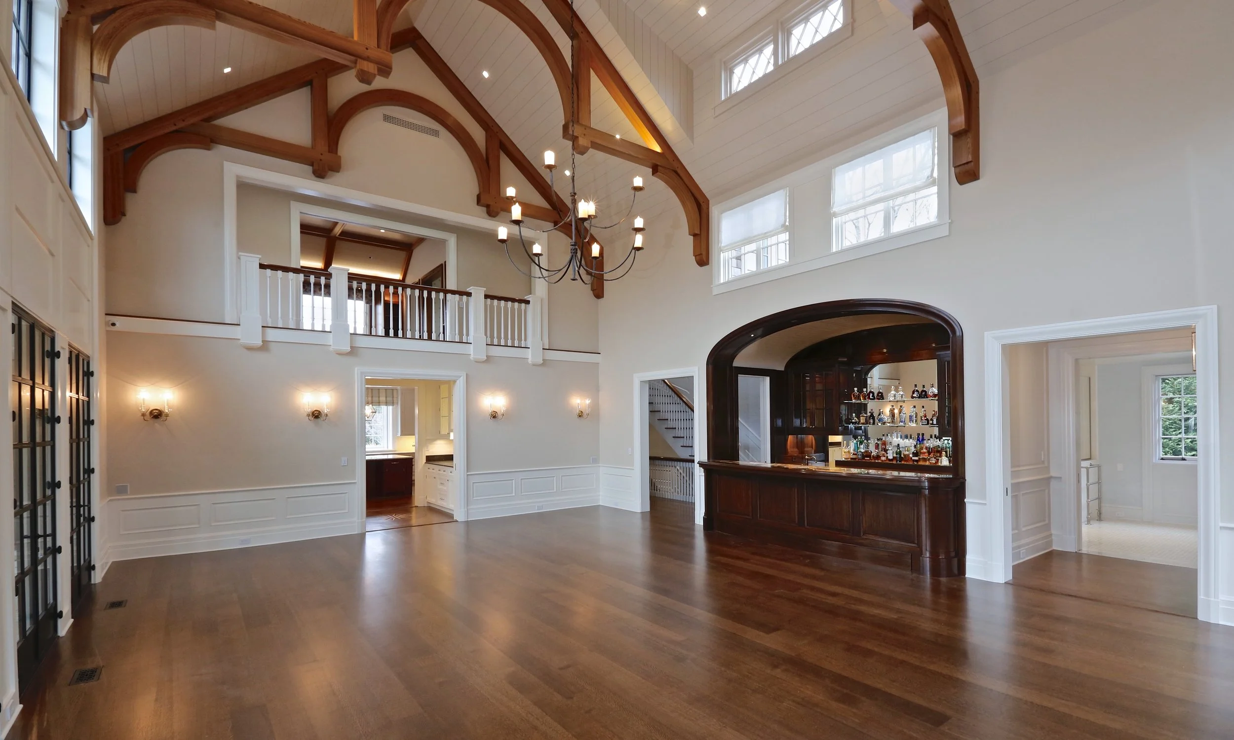 Spacious living room with high vaulted ceiling, wooden beams, chandelier, windows, and a built-in bar with bottles and glasses.
