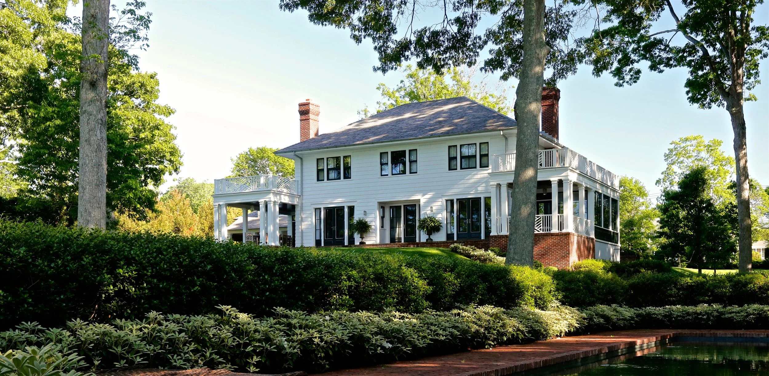 White two-story house with large porch, surrounded by greenery and tall trees, under a clear blue sky.