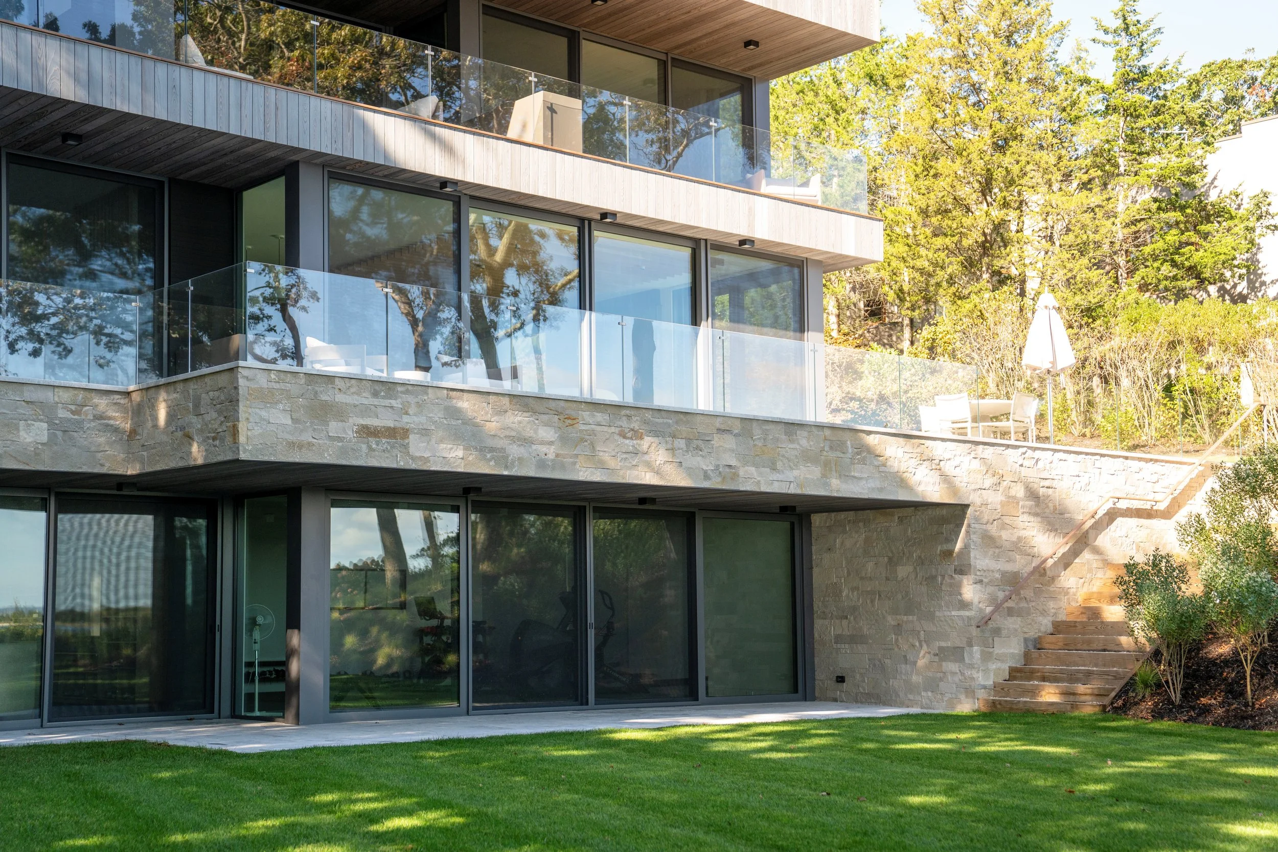 Modern multi-story house with large glass windows, stone wall accents, glass balcony railings, outdoor seating, and landscaped yard with stairs leading up to a terrace with patio furniture and umbrellas.