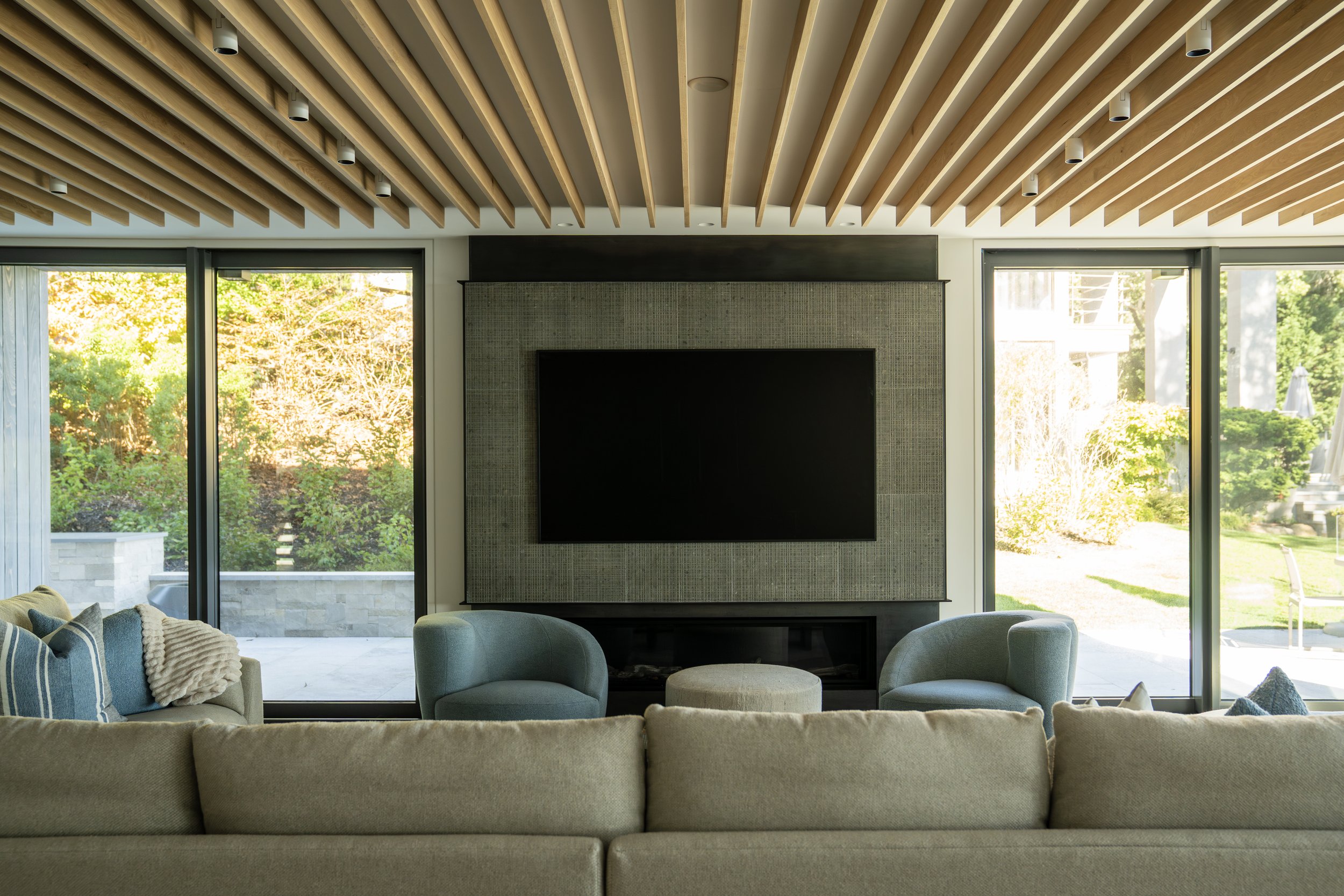 Modern living room with large windows, beige sofa, and two green armchairs facing a black TV mounted on a textured gray wall. A small round ottoman is centered between the chairs. Outside, lush green trees and a patio are visible.