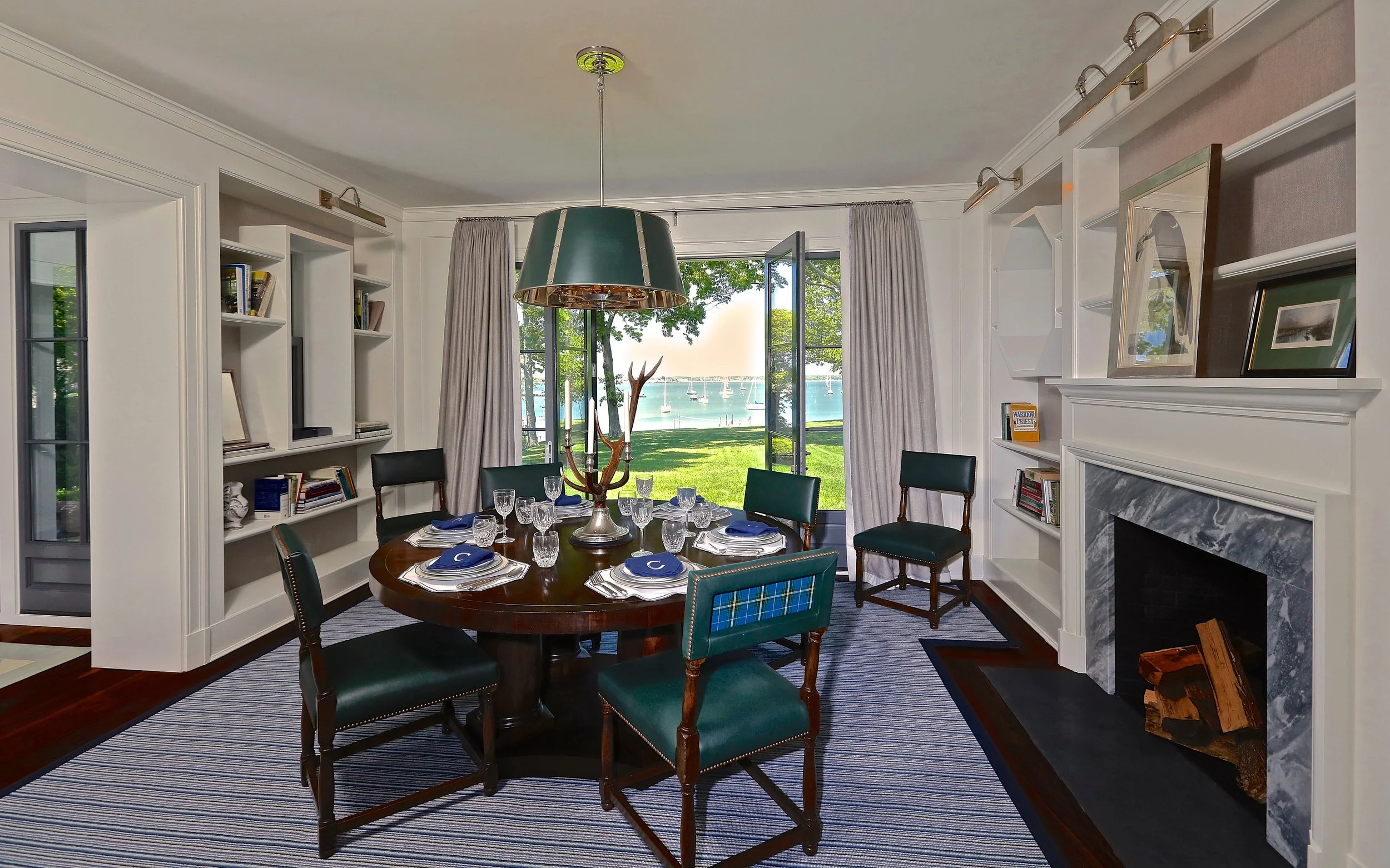 Dining room with a view of a lake, sailboats, and green trees through an open window, decorated with a round wooden table, green and plaid chairs, a chandelier, a bookshelf, and a marble fireplace.