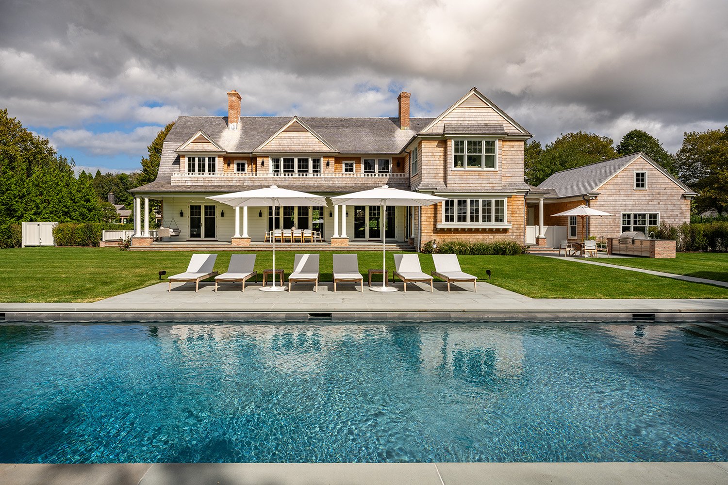 A large two-story house with a back porch, outdoor furniture, and a swimming pool with lounge chairs and umbrellas in the backyard.