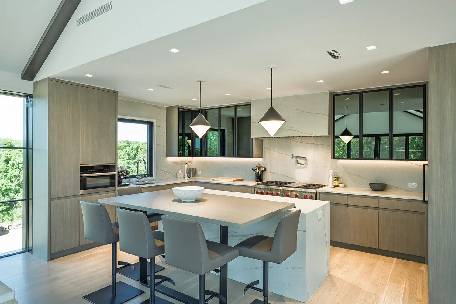 Modern kitchen with island, pendant lights, built-in oven, stove, and large windows with greenery outside