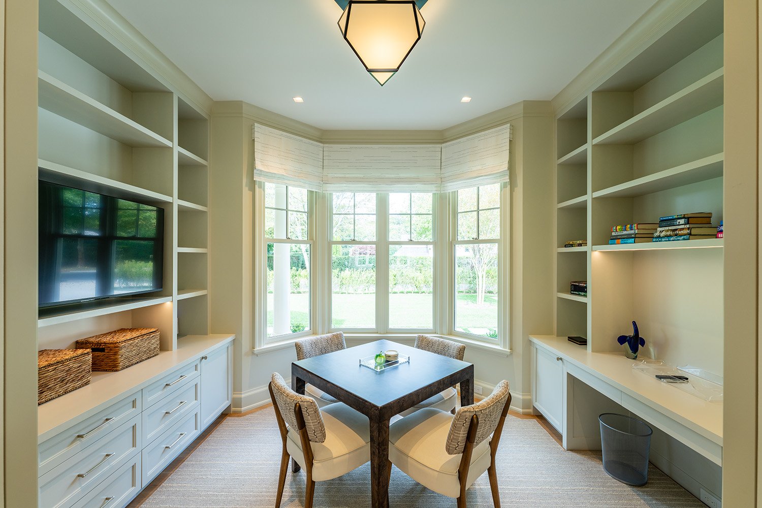 A bright dining area with a large bay window, a dark wood table, four beige chairs with patterned upholstery, white built-in shelves, a wall-mounted TV, and a ceiling light fixture.
