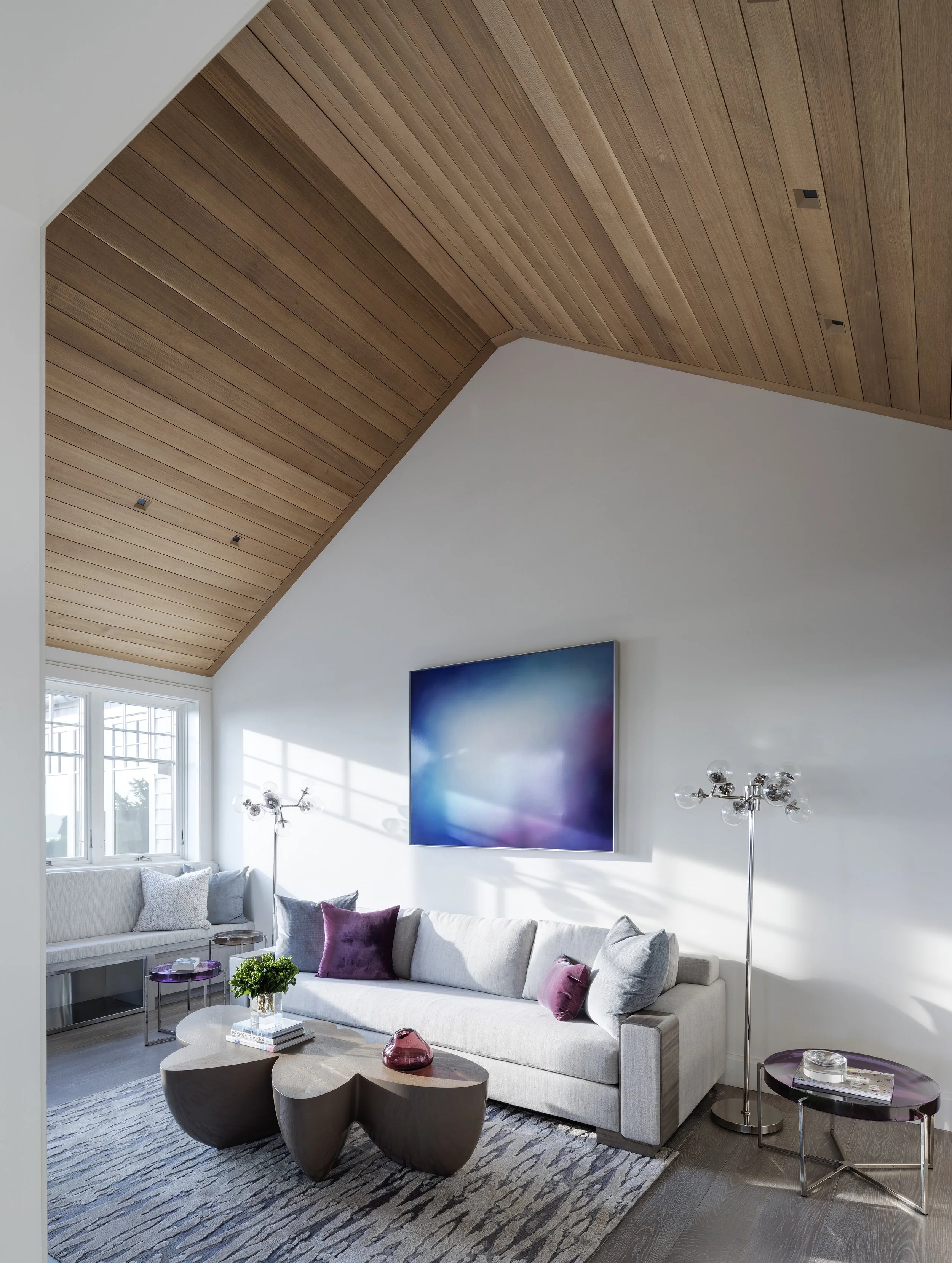Modern living room with a white sofa with purple and gray pillows, wooden coffee table, flat-screen TV on a white wall, and a window with sunlight, with a wooden ceiling.