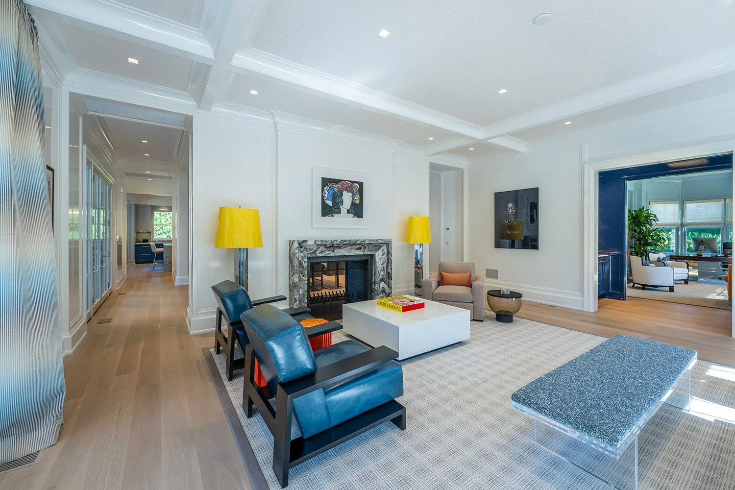 Living room with white walls, light hardwood floors, a marble fireplace, two yellow lamps, a small beige armchair, a white coffee table with a yellow and red book, three black armchairs, a bench with a sparkly blue cushion, and wall-mounted TV, with 