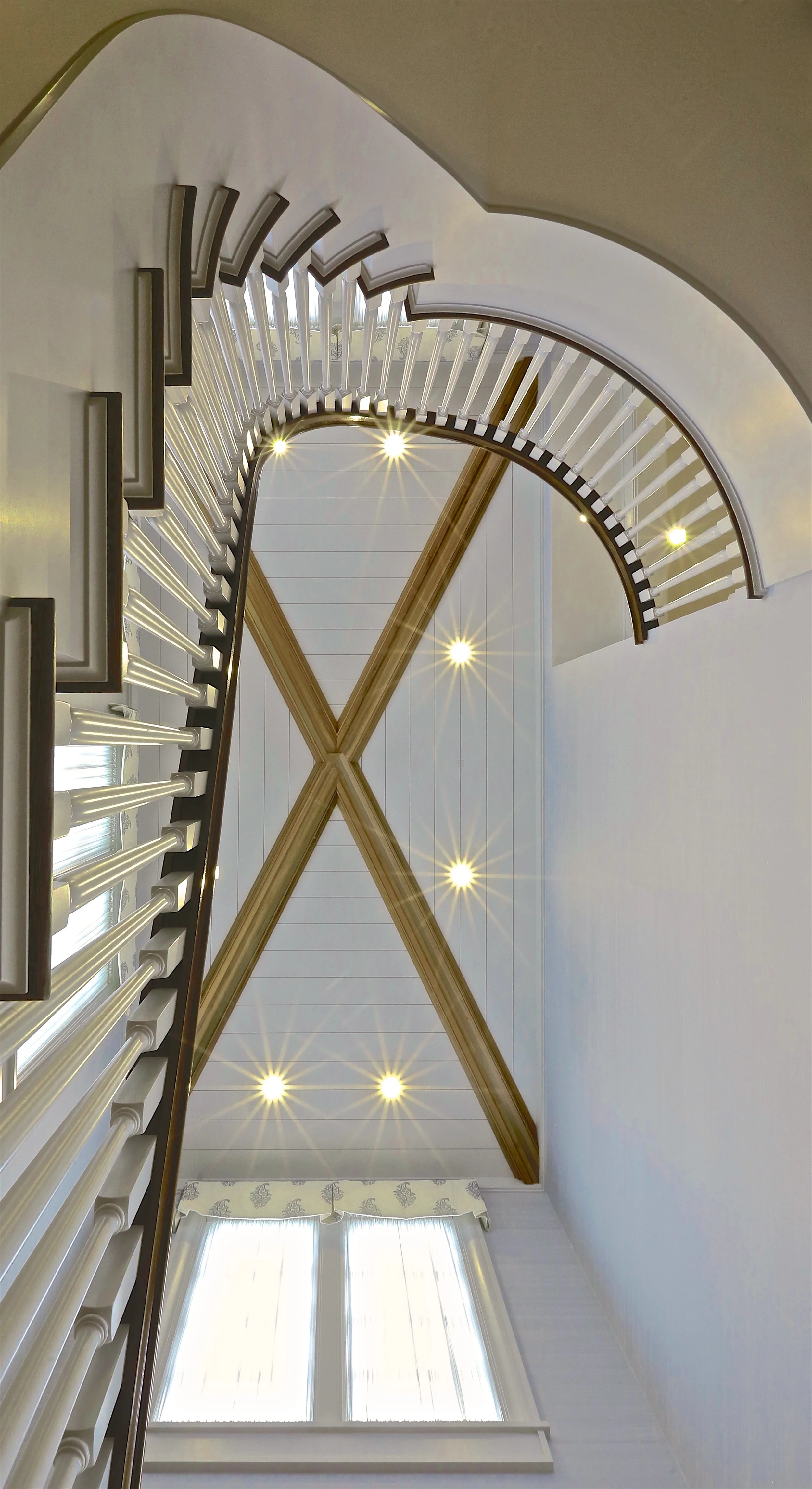 Looking up at a staircase and ceiling with wooden beams, recessed lighting, and a window with curtains.