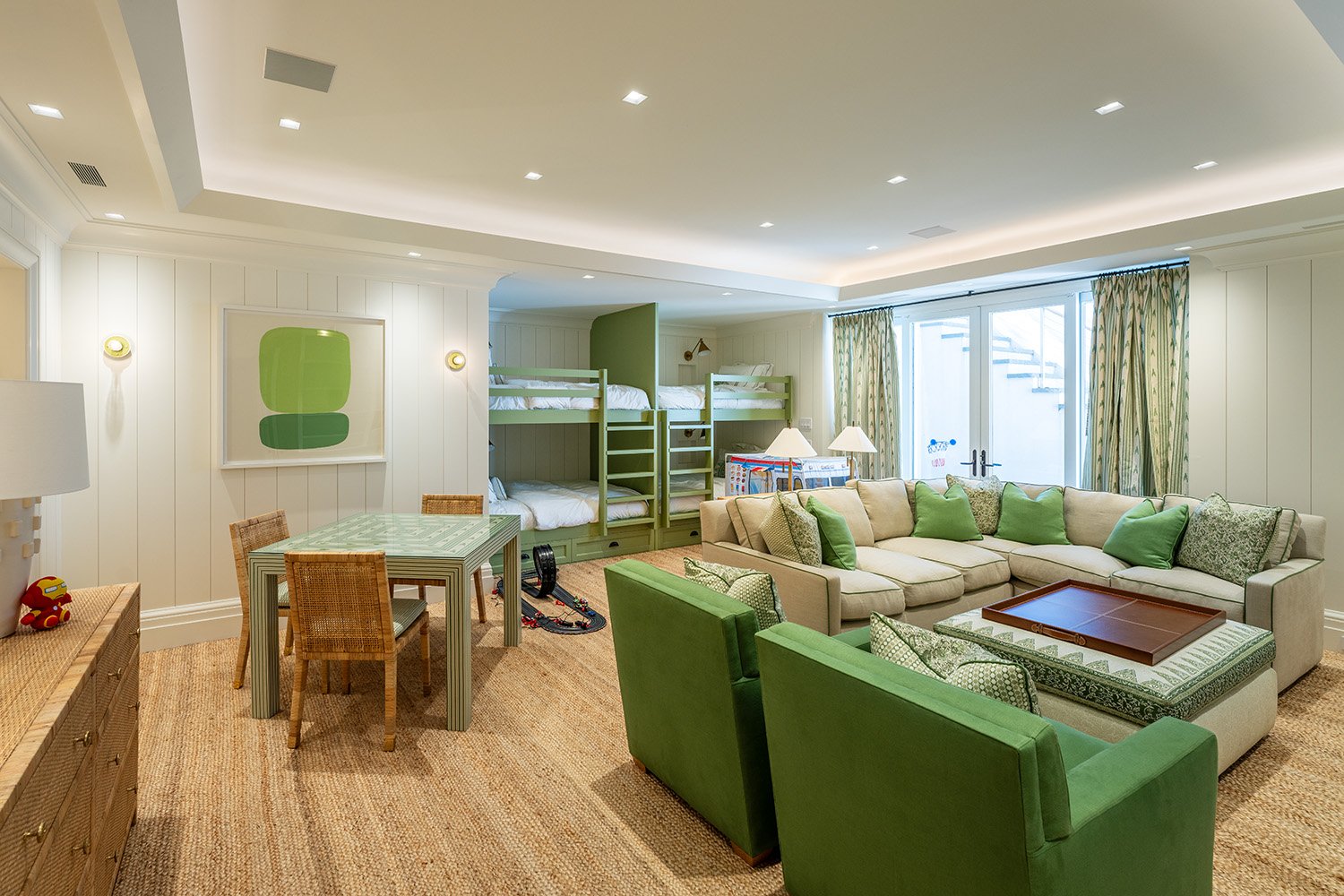 Large living room with loft beds, a beige sectional sofa with green throw pillows, green armchairs, a table with a tray, a wooden coffee table, a small dining table with four chairs, and large windows with patterned curtains.