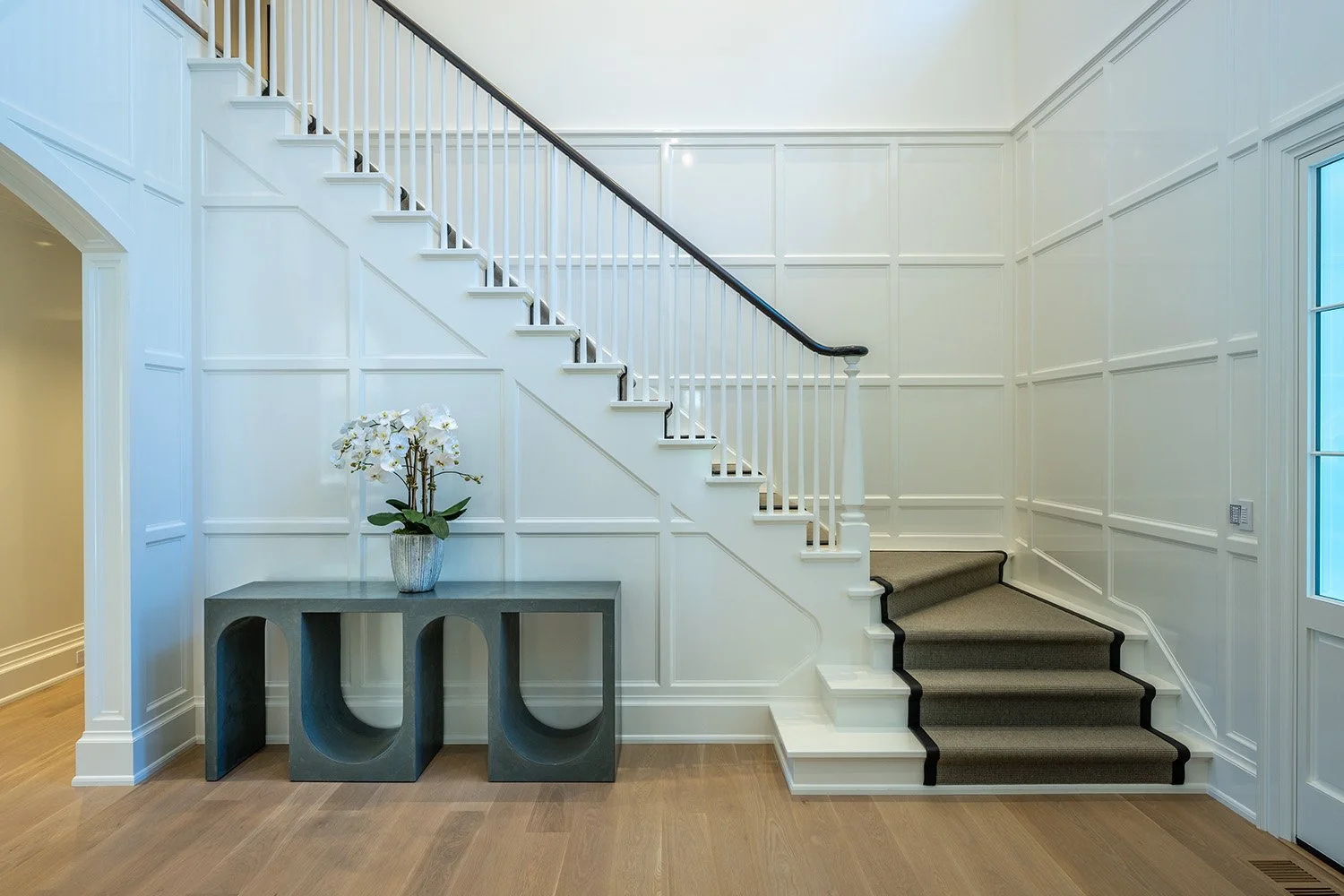 Interior view of a modern house entryway with white paneled walls, a staircase with wooden steps, and a black handrail, featuring a gray console table with a plant on it.