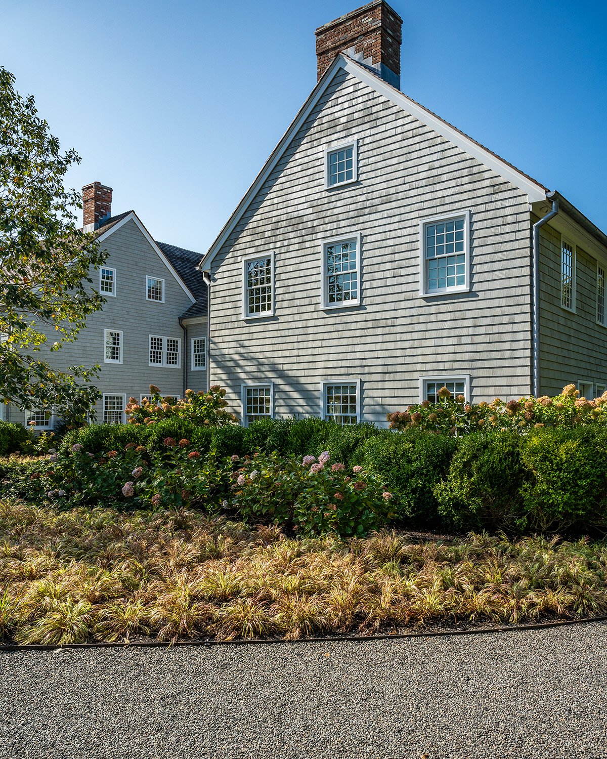 A white house with gray shingles, multiple windows, and brick chimneys, next to a garden with lush bushes, flowering plants, and a gravel pathway, under a clear blue sky.