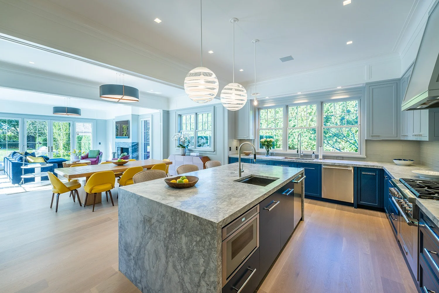 Modern open-concept kitchen and dining area with natural light, blue cabinets, marble countertops, yellow and beige dining chairs, and a living room with blue and pink furniture visible in the background.