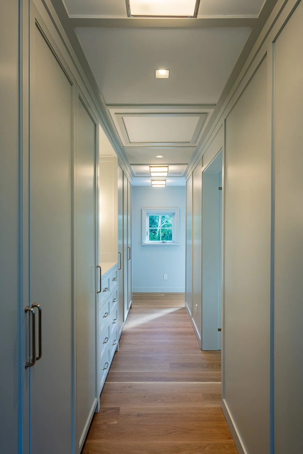 Empty hallway with white built-in cabinets on the left, a window at the end, wooden flooring, and ceiling lights.