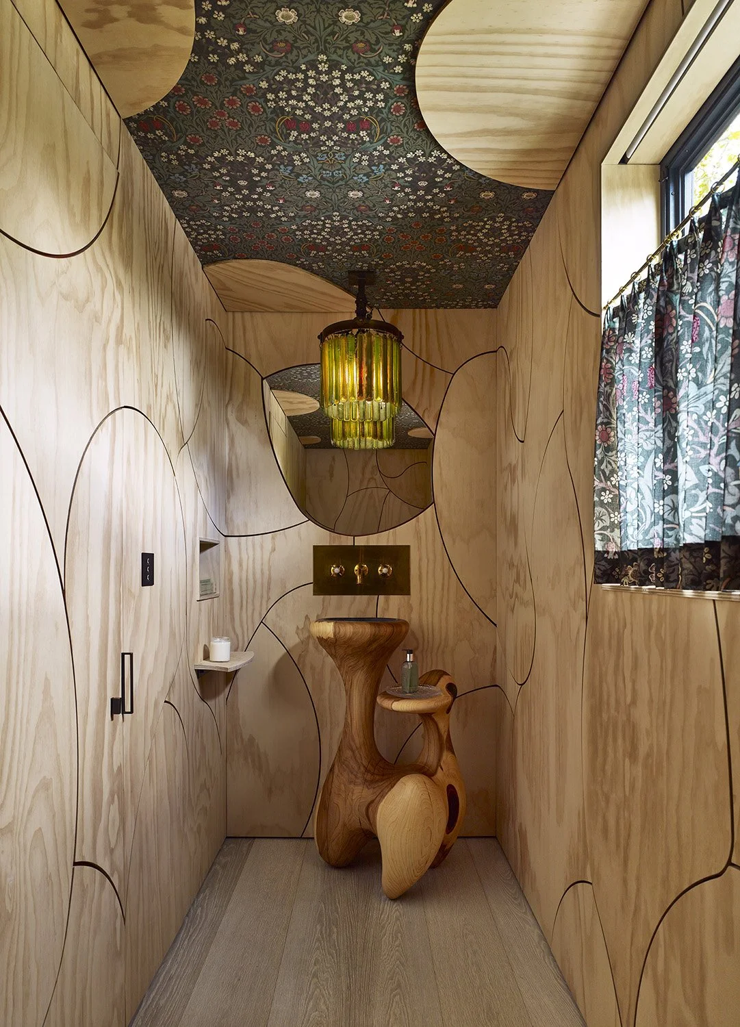Wood-paneled room with circular mirrors, patterned ceiling, a wooden sculpture table, a window with floral curtains, and a green hanging light.
