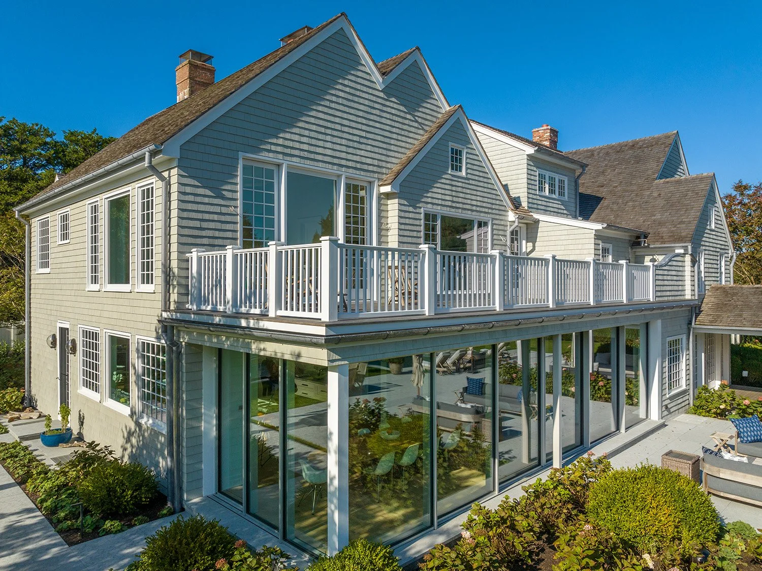 A large, multi-story house with gray siding, multiple windows, and a spacious balcony with a white railing. The house is surrounded by a well-maintained garden and has a glass-enclosed patio area on the ground floor.