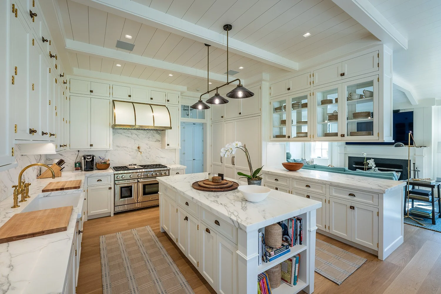 Modern kitchen with white cabinetry, marble countertops, and gold hardware. There is a central island with open shelving, a cooktop with a marble backsplash, and glass-front upper cabinets. Hardwood floors and pendant lighting complete the space.