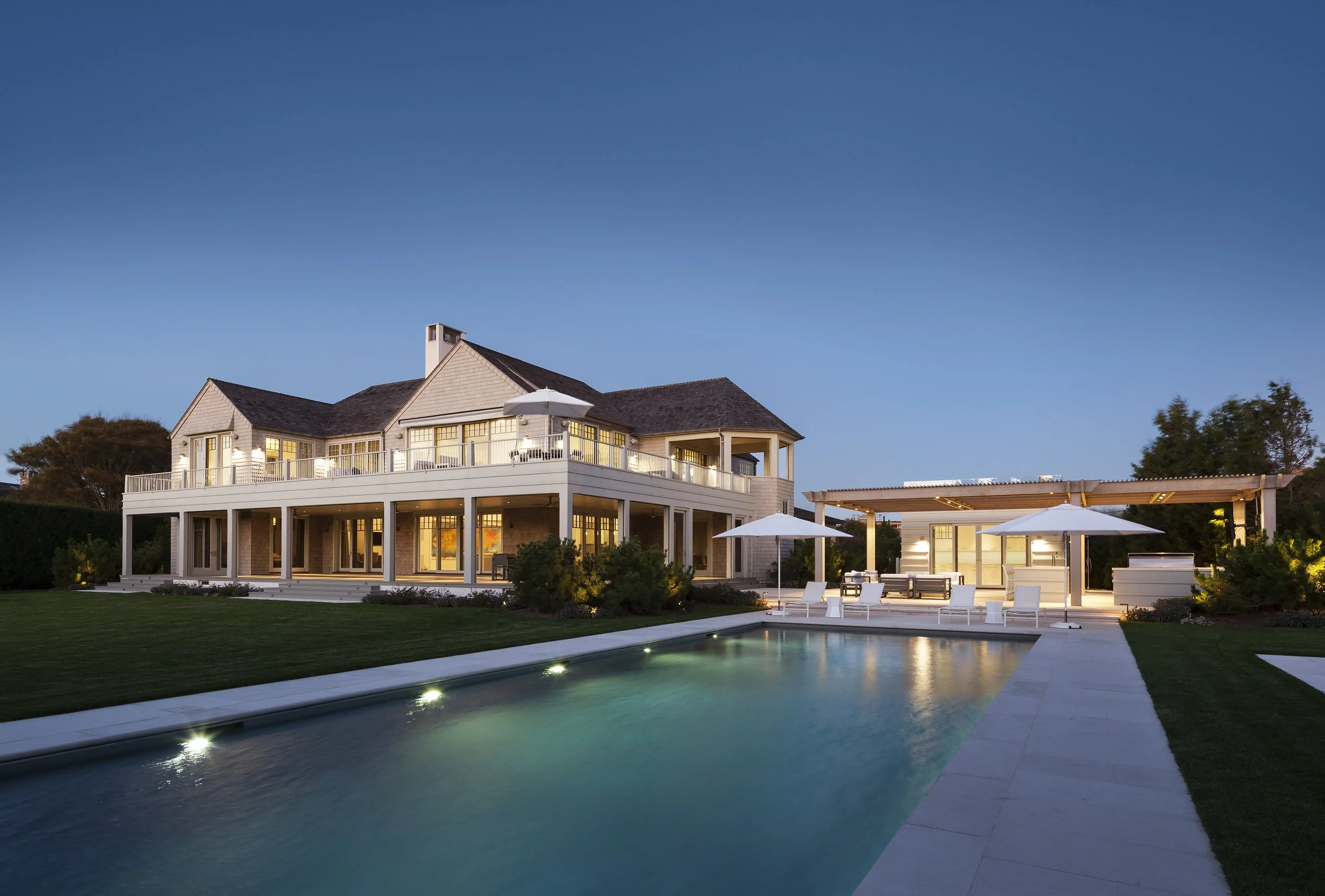 Luxury house with large deck, outdoor lounge, swimming pool, lawn, and evening lighting.