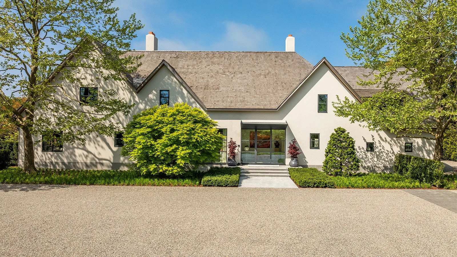 A modern house with a white exterior, a large front yard, and tall trees on both sides. The house has a gable roof and a glass front door with steps leading up to it. There are bushes and potted plants around the entrance.