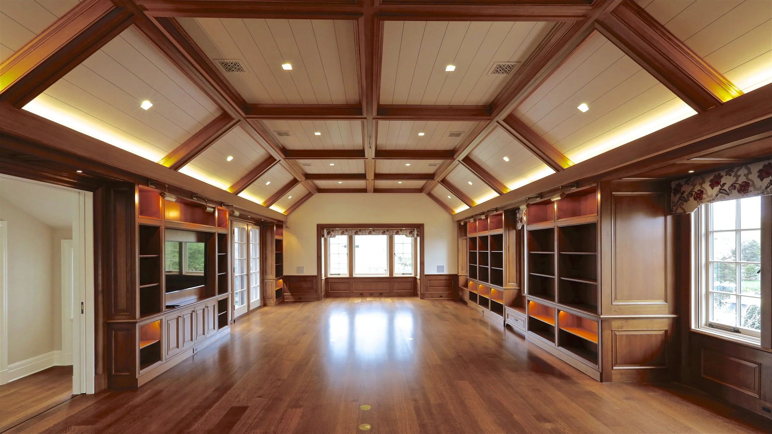 Large empty room with wooden flooring, wood-paneled walls, built-in bookshelves, and large windows, featuring a vaulted ceiling with wooden beams and recessed lighting.