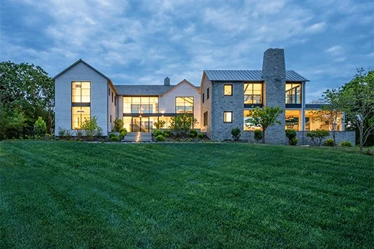 Modern house with large windows, stone chimney, and landscaped backyard at dusk.