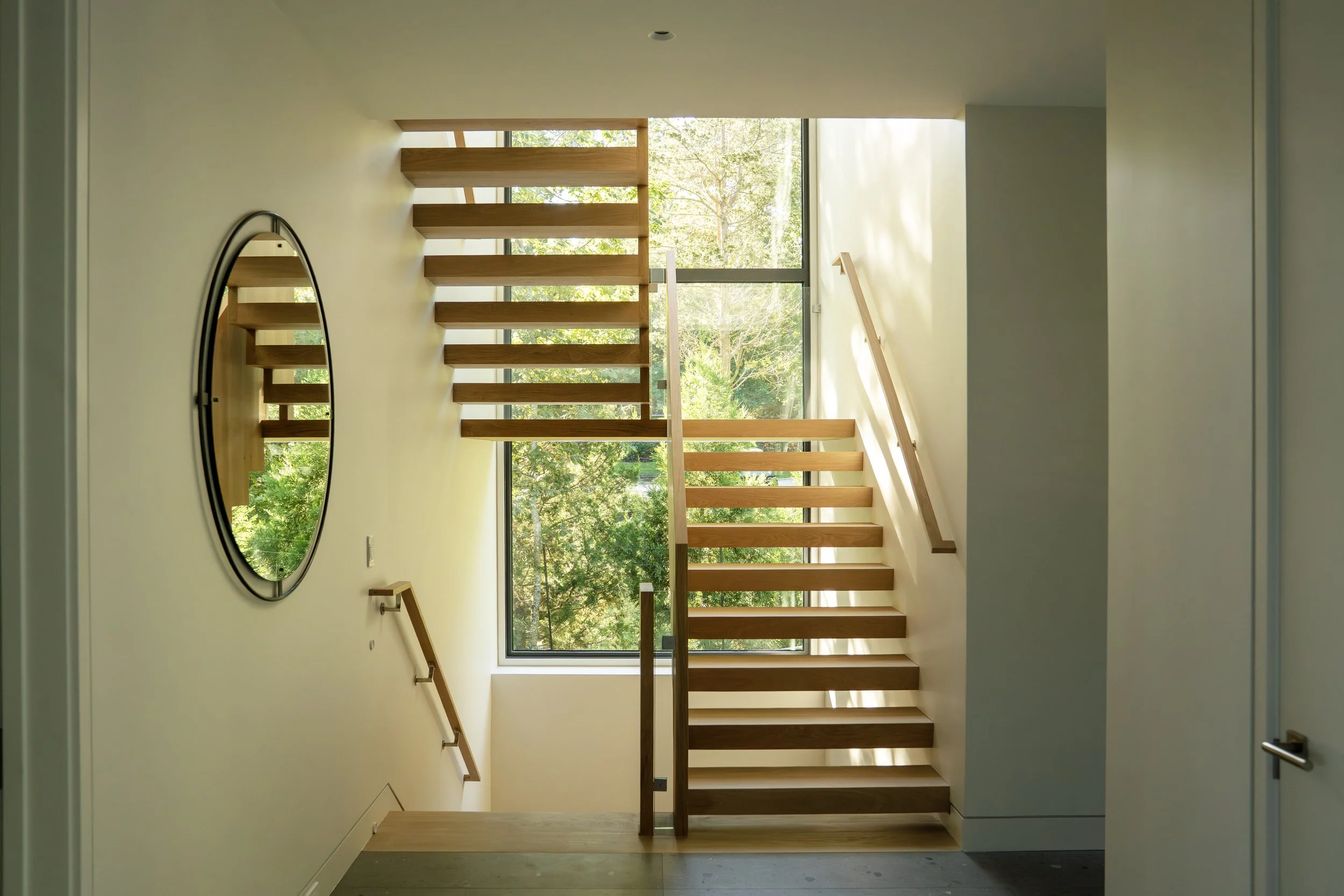 Modern indoor staircase with wooden steps and a large window showing trees outside, with a round mirror on the wall.