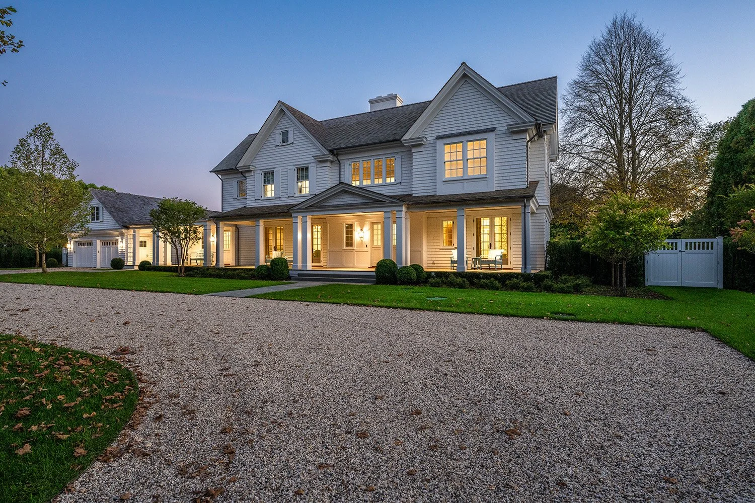 Large white two-story house with front porch, illuminated windows, surrounded by a well-kept lawn, trees, and a gravel driveway at dusk.