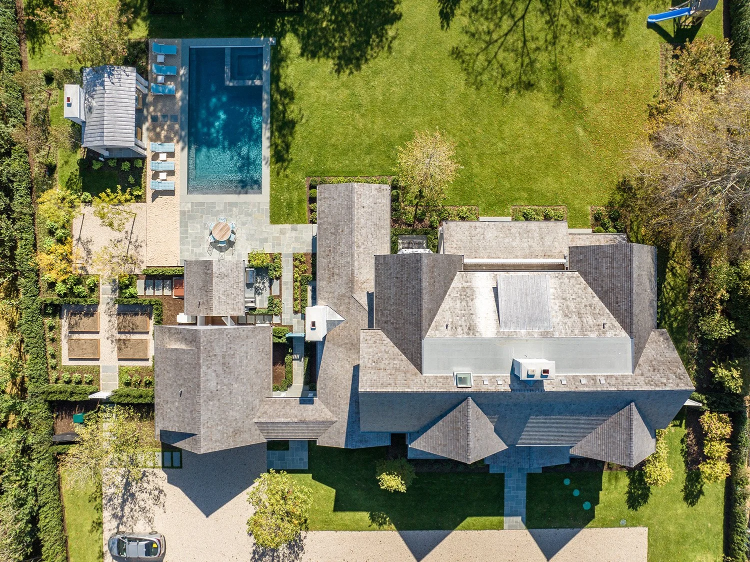 Aerial view of a residential property showing a large house with various roof sections, a backyard with a swimming pool, patio area with outdoor furniture, and surrounding greenery including trees, shrubs, and a well-manicured lawn.