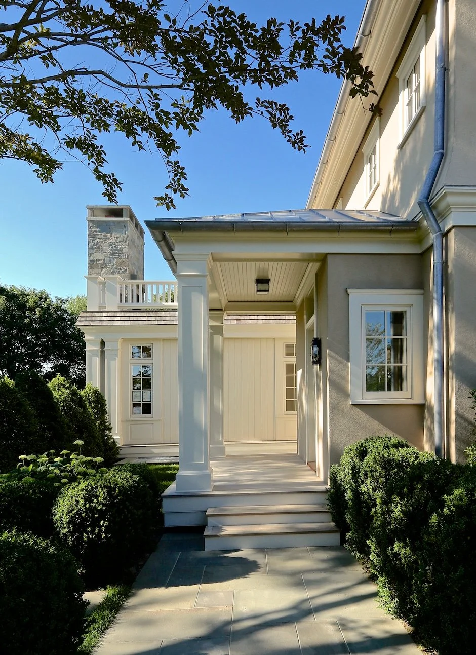 Front view of a modern house with a porch, columns, a stone chimney, and lush green bushes under a clear blue sky. Sag Harbor construction