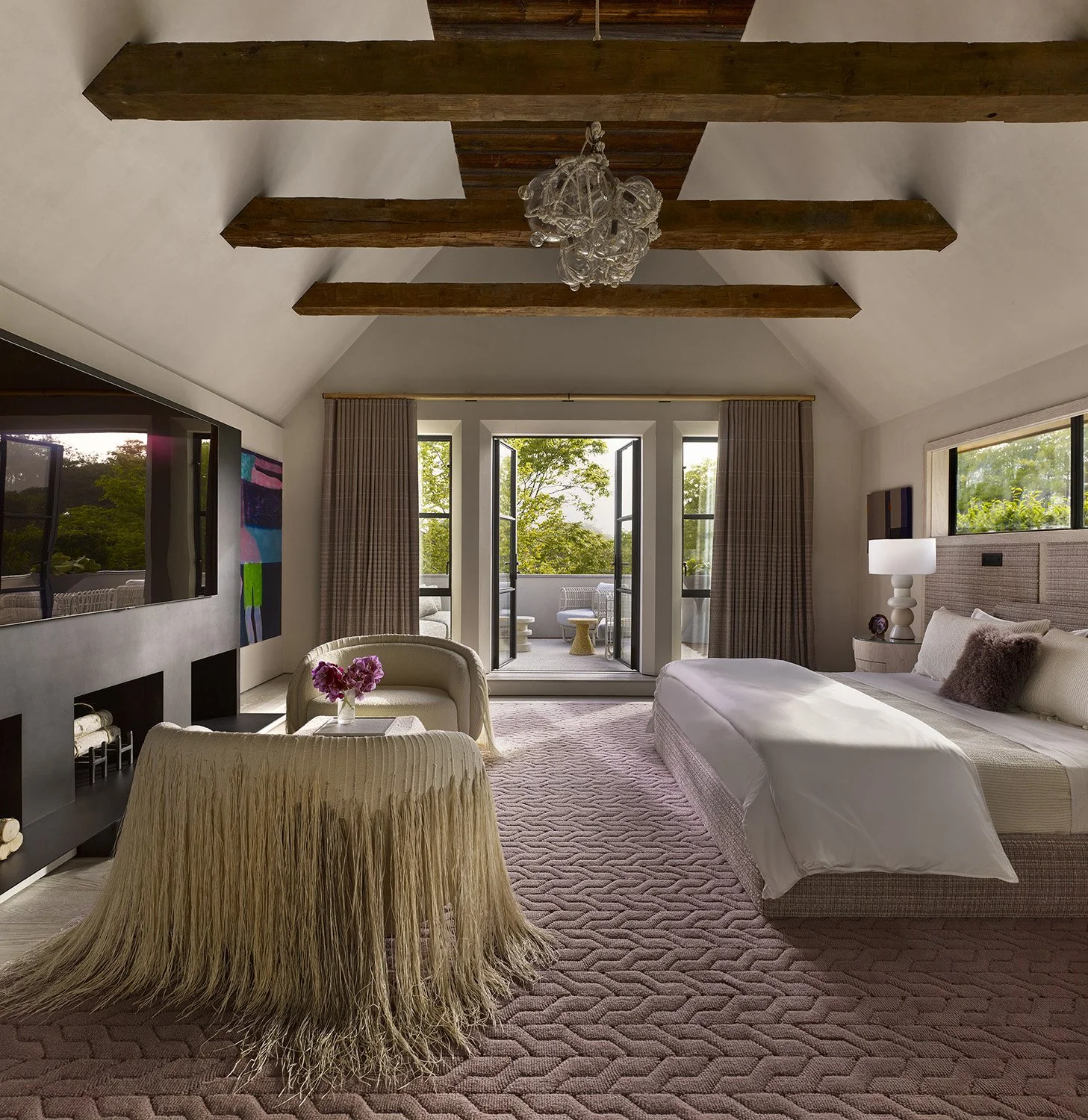 A bedroom with a vaulted ceiling, wooden beams, and a glass chandelier. There is a large bed with white linens and pillows, a bedside lamp, and a small seating area with a curved sofa and a flower arrangement. Large windows and a balcony door with cu