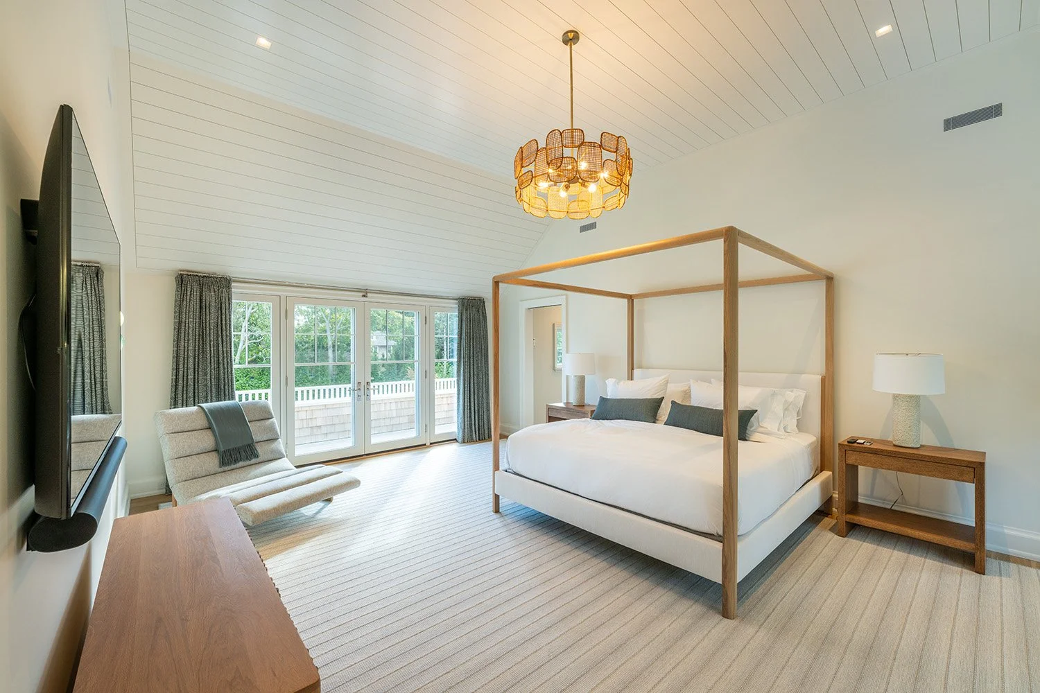 Bright hotel bedroom with a four-poster bed, nightstands, table lamps, a lounge chair, and glass doors leading to a balcony with a view of trees.