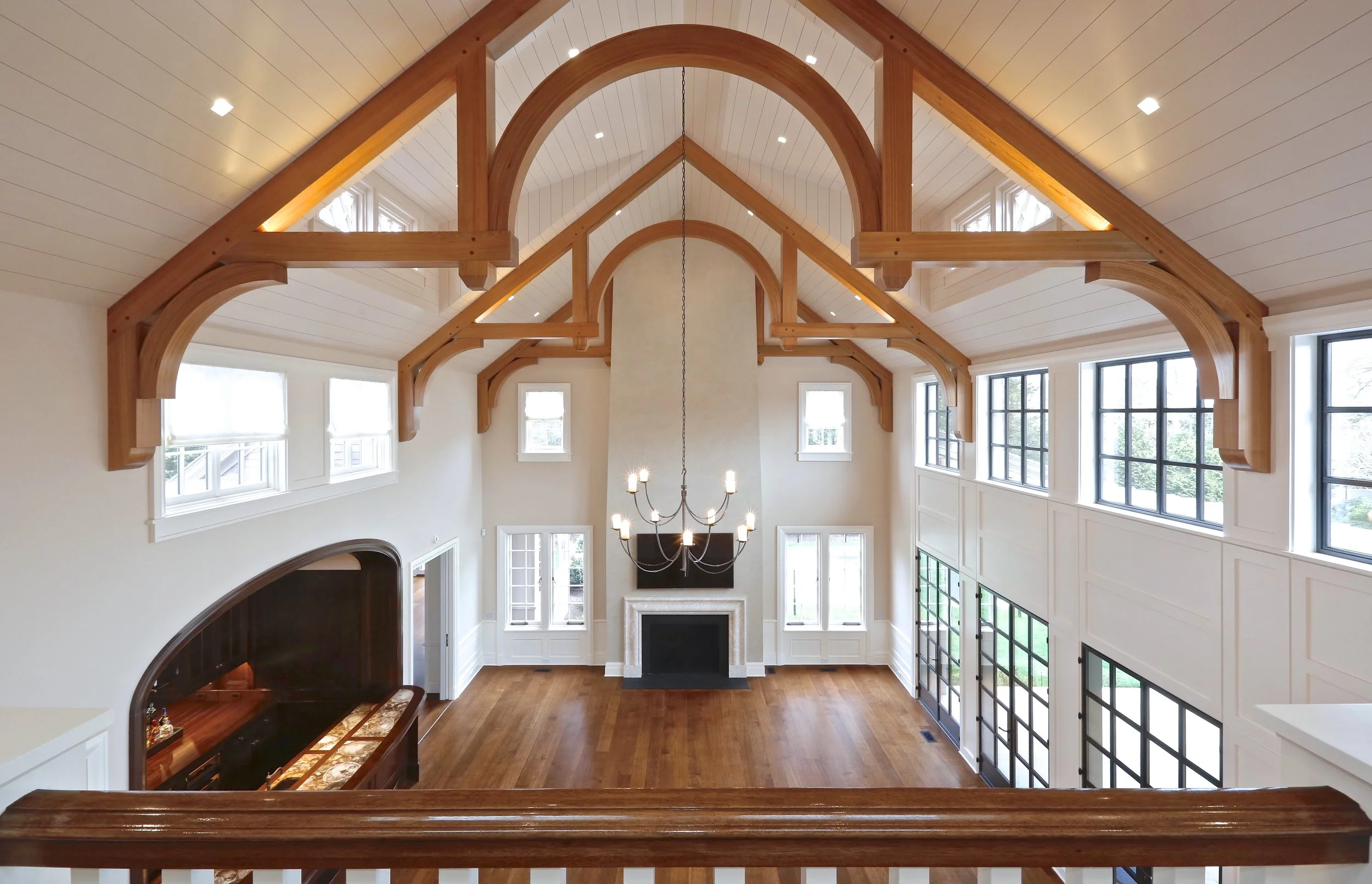 Interior view of a spacious living room with high vaulted ceilings, wooden beams, large windows, a fireplace, and a chandelier.