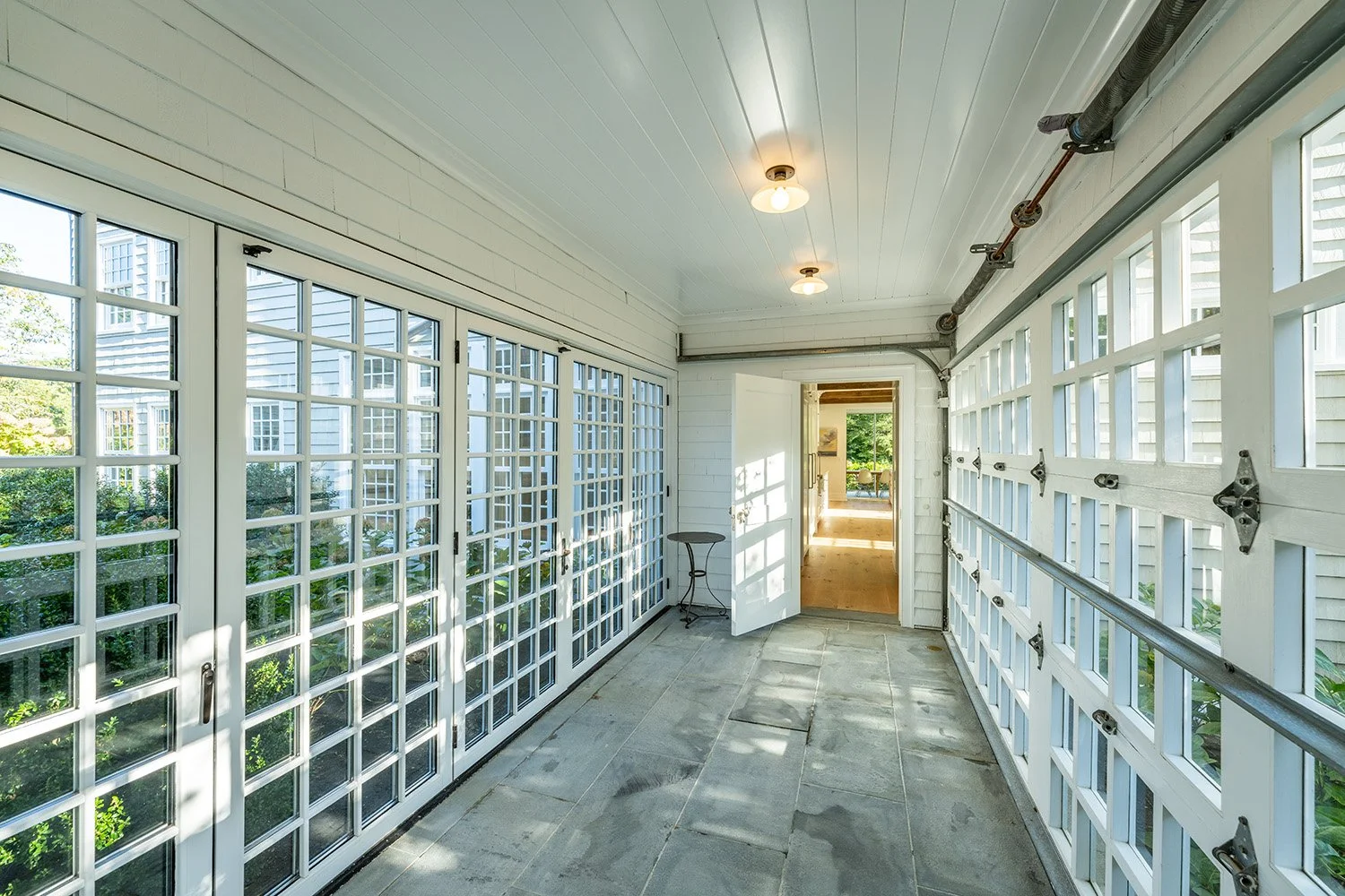 A sunlit enclosed porch with white framed glass doors and windows, gray stone flooring, and an open door leading to an interior room with wooden floors and greenery outside.