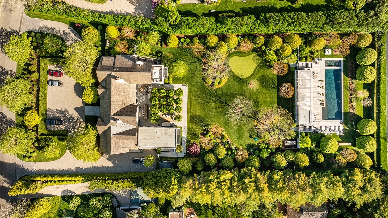 Aerial view of a large residential backyard with a house, parking area, lush trees, a small golf putting green, a swim pool, and a deck area.