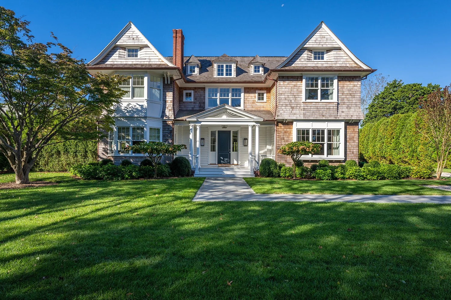 A large, multi-story house with a front porch, surrounded by a well-maintained lawn and green trees, under a clear blue sky.