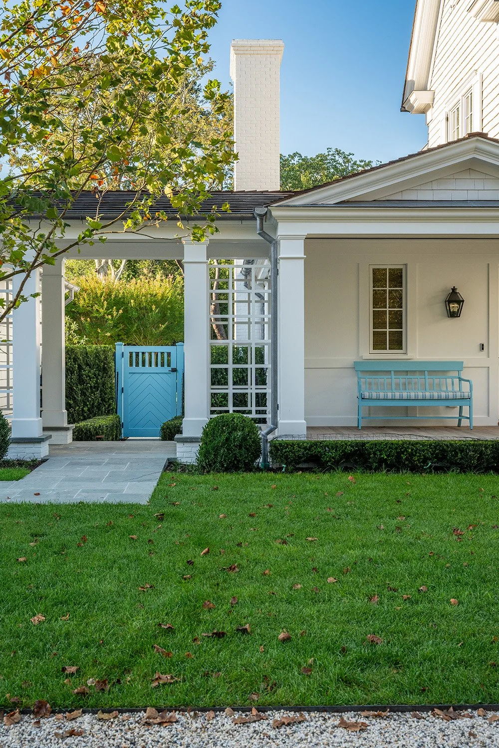 Front porch of a white house with a light blue bench, a small window, and a black outdoor lantern. A light blue gate leads to a backyard with green shrubs and trees, and a paved pathway. The yard has a well-maintained green lawn.