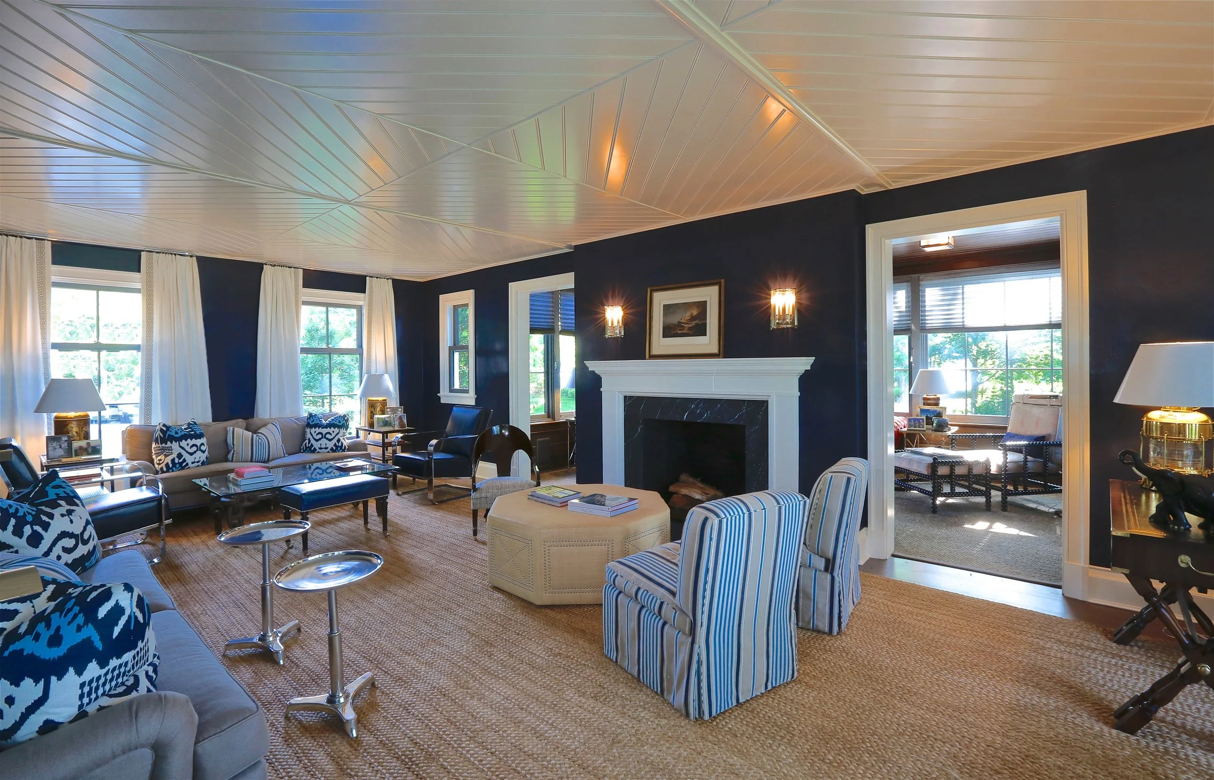 A spacious living room with navy blue walls, white curtains, and multiple windows letting in natural light. The room features a large fireplace with a white mantel, surrounded by assorted chairs including striped and solid-colored ones, a sofa with p