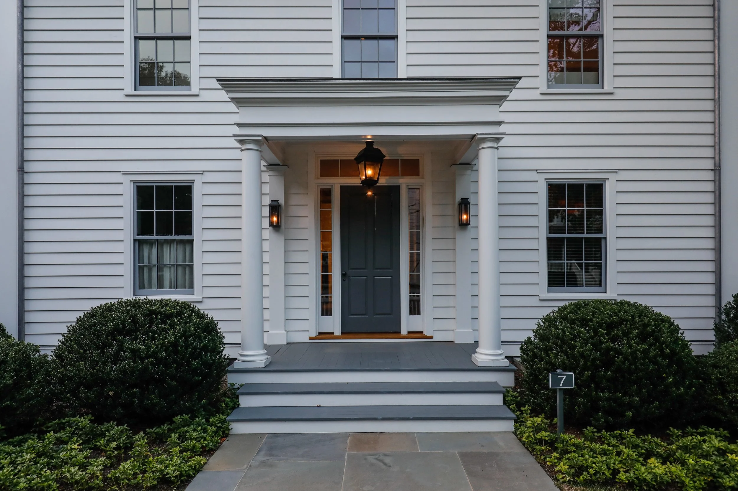 Front view of a white house with a covered porch, grey front door, and two windows on each side. The porch has two columns, steps leading up to it, and a hanging black lantern with two wall-mounted lanterns on either side of the door. There are well-