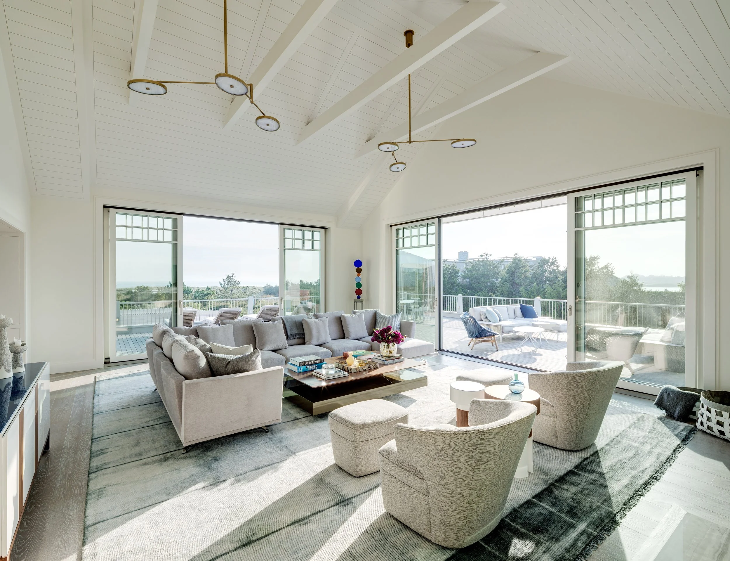 Bright living room with large sliding glass doors leading to outdoor patio, featuring a sectional sofa, armchairs, coffee tables, and modern ceiling lights.