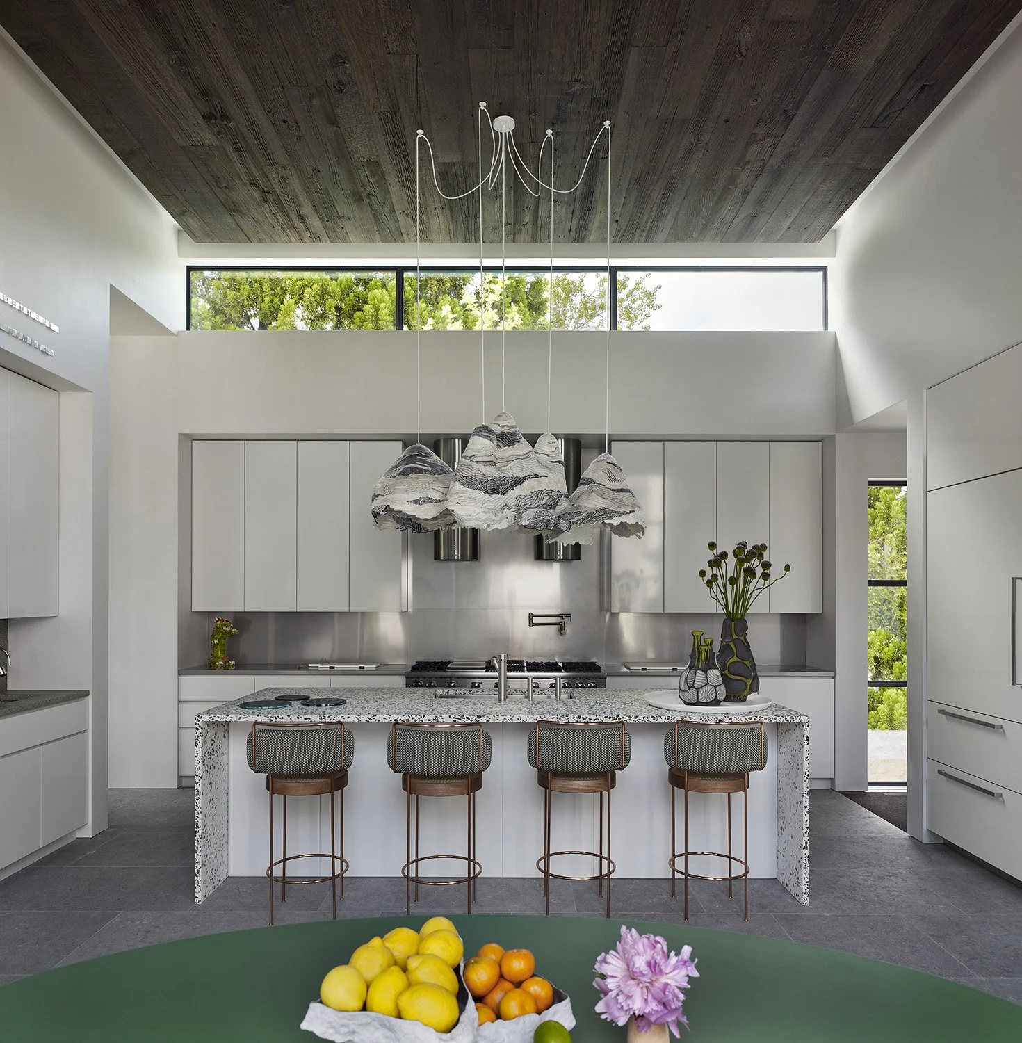 Modern kitchen with white cabinets, marble island, and artistic pendant lights casting cloud shapes, with large windows and greenery outside.