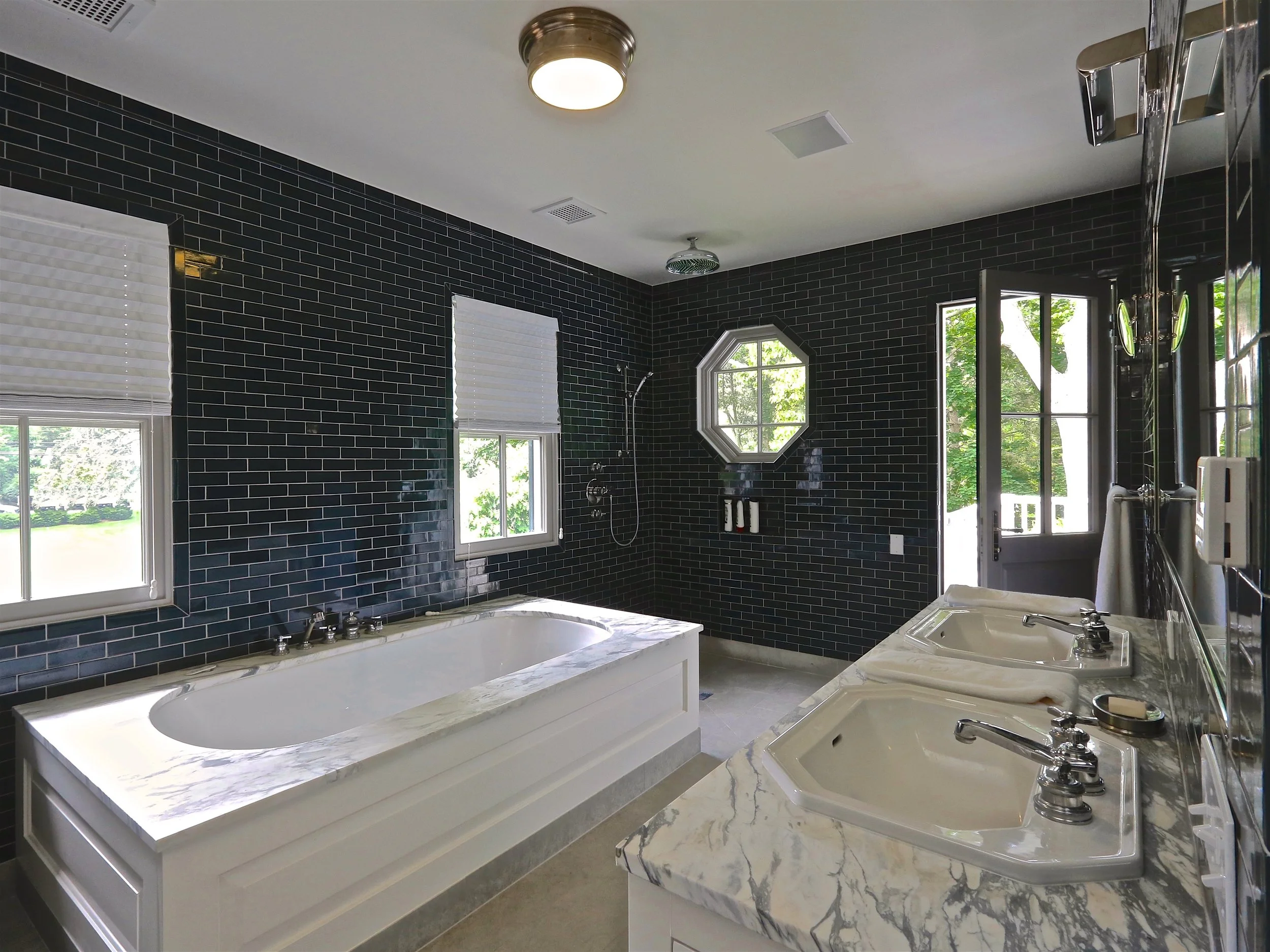 Bathroom with dark blue subway tile walls, a built-in bathtub with a marble surround, double sinks with marble countertops, two windows with white blinds, a shower with a rain head, and a door leading outside, with greenery visible through the window