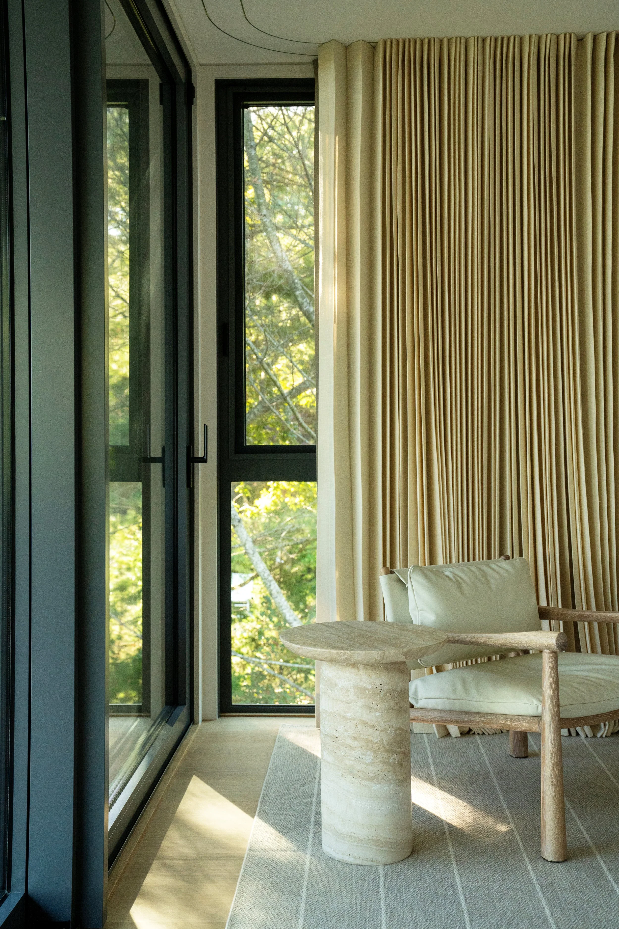 A corner of a modern living room featuring a large window with a view of trees, beige curtains, a wooden chair with cream cushions, a round stone side table, and a light-colored area rug.
