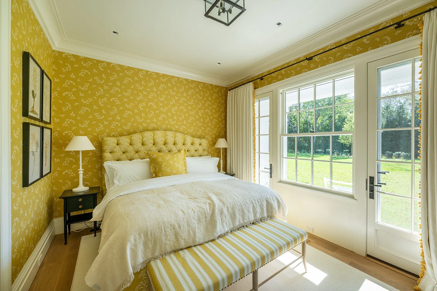Bedroom with yellow floral wallpaper, large bed with tufted headboard, white bedding, yellow pillows, two bedside lamps, large window with white curtains, outside view of green yard with trees.