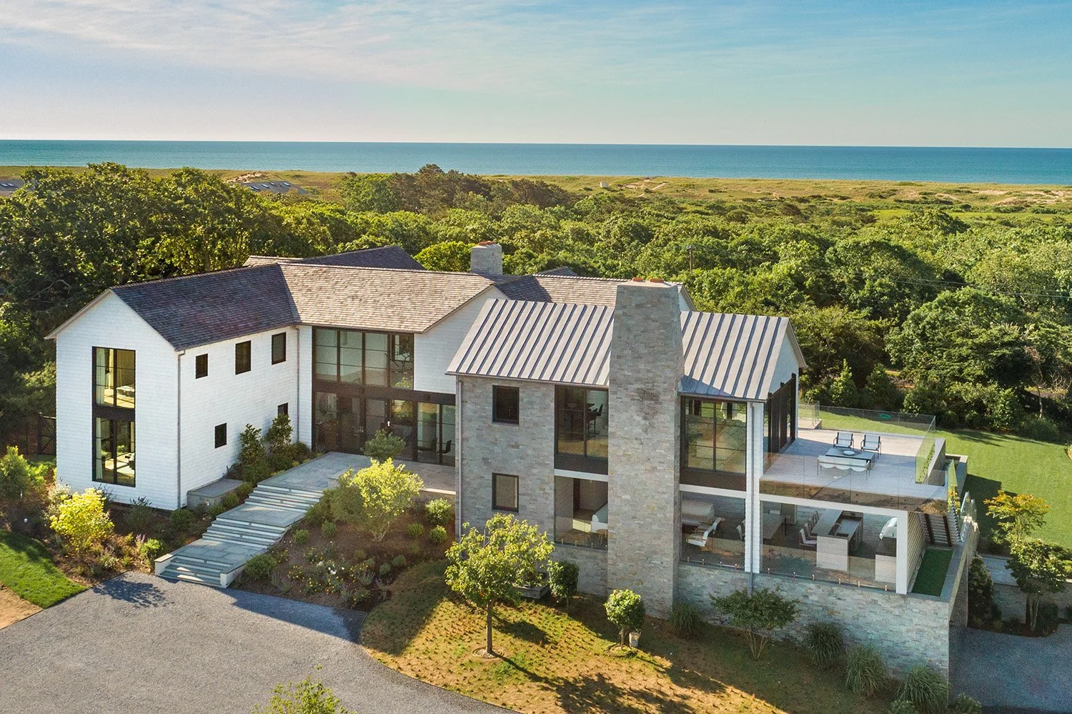 A modern, multi-story house with a mix of white and brick exterior, large windows, surrounded by greenery, with a view of the ocean in the background.