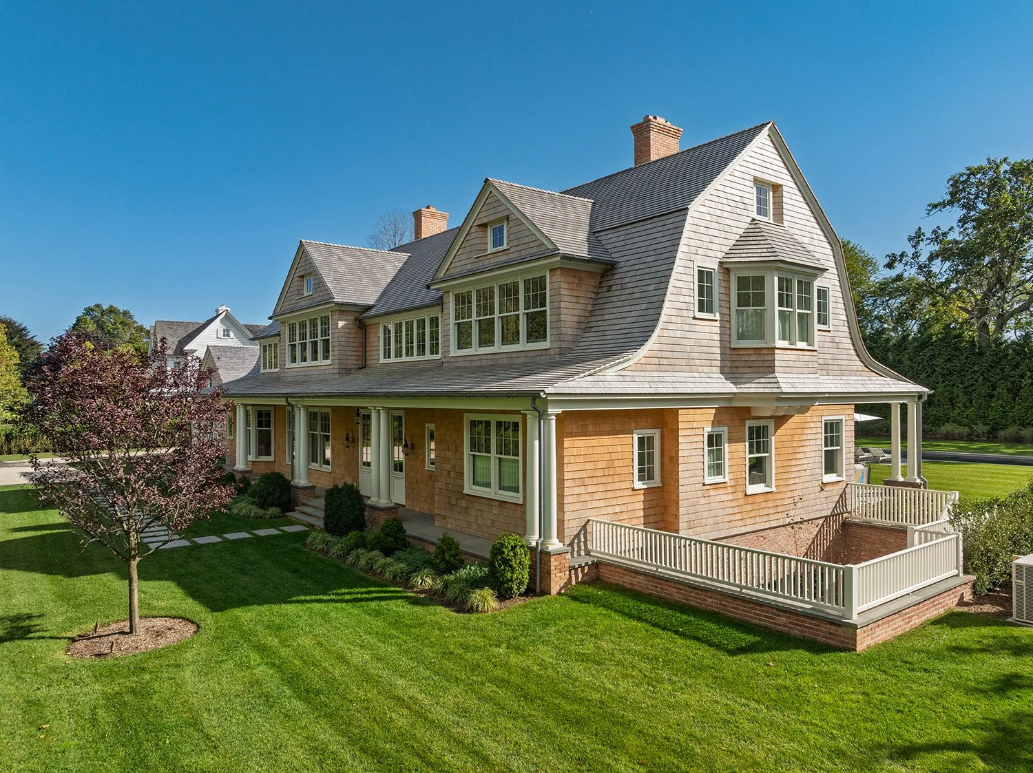 A large historic-style house with multiple gabled roofs, shingle siding, a wraparound porch with columns, and a landscaped front yard with a small flowering tree and green lawn. Construction in Hamptons New York