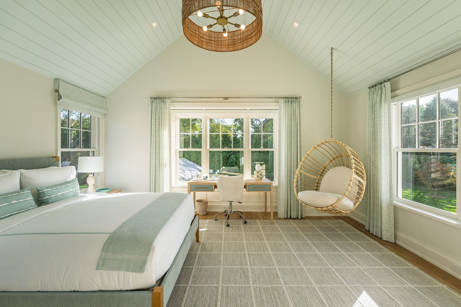 Bright bedroom with white walls, a large bed with green accent pillows, a desk by the window with a white chair, and an empty hanging wicker chair.