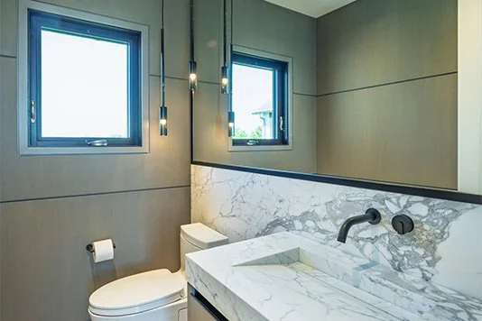 Modern bathroom with gray walls, two small windows, a white toilet, a marble sink, and black fixtures.