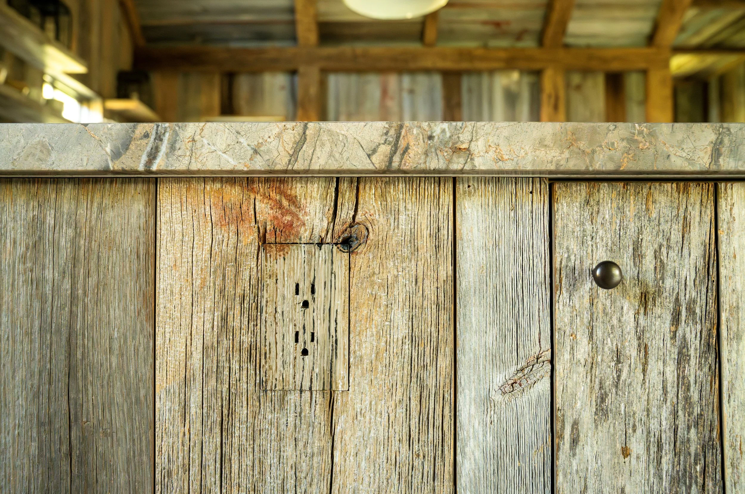 Close-up of rustic wooden paneling with weathered textures and visible knots, topped with a stone countertop, inside a barn or rustic kitchen.